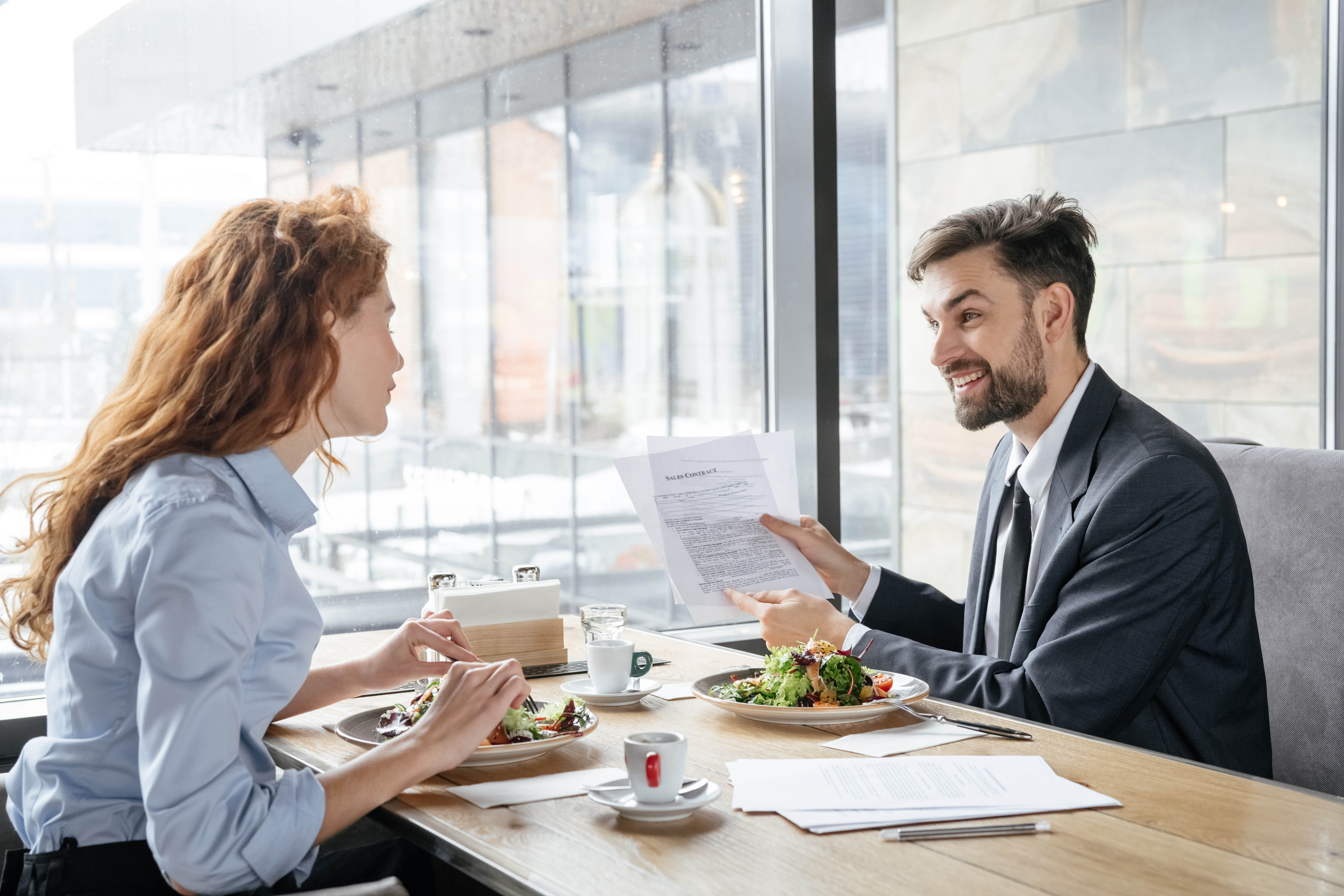 Young man and woman are having a business lunch and reviewing papers that he's holding up, while eating their salads Young man and woman are having a business lunch and reviewing papers that he's holding up, while eating their salads